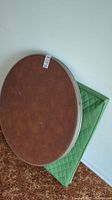 Top view of one brown wood-tone vintage folding card table leaning against a green padded case against a white wall and brown carpet.