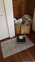 Wicker baskets with white plastic clothes hangers inside and around, basket tote with green handles, all positioned on floor near white cabinet and wooden wall. Gray bathmat underneath the Taylor bathroom scale.