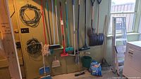 Wall-mounted pegboard displaying brooms, mops, dustpans, shovel, rake, bucket, extension cords, and a lightweight step ladder near a window.