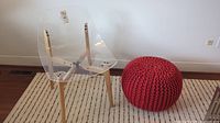 Full view of clear plastic chair with wooden legs and red knit pouf on a patterned rug