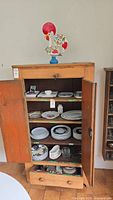 Wooden cabinet with open doors showing various plates and bowls on shelves, along with a colorful rooster decoration on top.