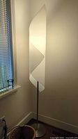 Photo of a tall white floor lamp with a wavy modern design shade and metal stand placed near a wall and window.