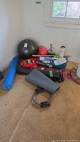 Wide shot of exercise equipment cluster including foam roller, Pilates ring, yoga mats, kettlebell, dumbbells, and stability ball on carpeted floor near wall and window.