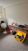 Room corner with two electric fans, a yellow toolbox on the carpet, and hardware items on a brick ledge.