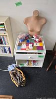 Wide view showing white shelf with drawer containing yarn, knitting projects, books, and the headless mannequin behind it. Knitting basket with needles on the floor.