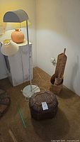 Floor lamp with white curved shade, table lamp with terracotta base, round woven basket with lid, and tall woven basket with pointed lid and two small cylindrical baskets next to it on carpeted floor against white wall.