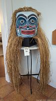 Full view of painted styrofoam mask with natural fiber hair on black metal-legged table with marble top, showing length and proportions.