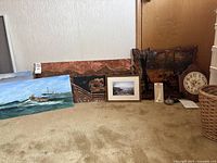 Overall lot contents arranged on floor showing paintings, copper artwork, clocks, pillow, small decorative item and basket.