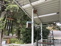 Two wooden bird houses hanging from a white patio roof with one featuring black wind chimes, surrounded by outdoor greenery.