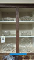 Front view of wooden cabinet shelf with multiple crystal and glass serving dishes displayed, showing various patterns and bowl shapes.