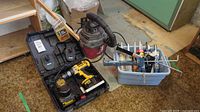 Wide view showing DeWalt cordless drill in case, Shop-Vac, and gray bin with caulking items on floor.