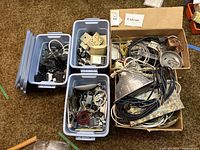 Wide view of three plastic bins and one cardboard box filled with various electrical components including switches, sockets, wiring, metal parts, and plugs