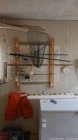 Photo of fishing poles mounted on a wooden rack with fishing net and orange life jacket below them, along with rolled maps visible.