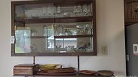 Wall-mounted wooden cabinet with glass doors holding an assortment of clear glassware, including wine glasses, tumblers, and bowls. Below the cabinet on a flat surface are wooden serving trays and a wooden box.