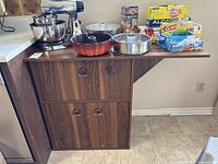 Front view of brown MDF kitchen or bar cabinet with fold-down side table loaded with kitchen items.