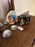 Black lacquer Japanese themed jewelry music box, decorative Florida paper fan, and two floral tea cups with saucers on wood table.