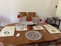 Wooden table with a pink glass cake stand, pinwheel crystal platter, metal fish platter, four red candle holders, and six bird-themed placemats arranged on it.