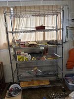 Full view of shelving unit with multiple shelves holding various small items and tools, in front of a window with curtains.