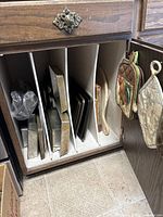 Inside cabinet showing multiple vertically stored baking sheets and pans, small cutting board, two oven mitts hanging on cabinet door