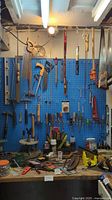 Photo showing variety of saws, levels, hand clamp, and hanging tools on a blue pegboard wall above a cluttered workbench with various small tools and materials.