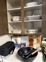 Kitchen cabinet shelves with multiple Corning Ware and stoneware serving dishes, white ceramic casserole dishes, black large ribbed bowl with utensils, Starfrit mandoline box, Tupperware containers, and a black soft case bag.