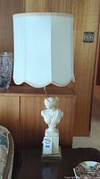 Full view of the vintage Artemis sculpture lamp with white scalloped-edge fabric lampshade on wooden side table, showing the lamp's base and shade against wood panel background.