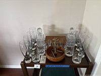 Wide view of wooden table showing vintage wooden cigar box on top, two crystal decanters with stoppers on either side, seven clear crystal pint glasses with cut pattern arranged around them, and wooden bowl with cocktail picks in center.