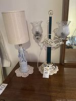 Photo showing two lamps: one Blue Boy figurine lamp with beige fabric shade on left, green glass double shade lamp with frosted scalloped glass shades on right, both on wooden desk.
