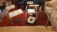 Full view of the lot showing walker with basket, white shower seat, commode with broken hinge and cushions on red carpet.
