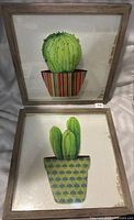 Two 14x14 framed cactus prints, one with a round cactus in a striped pot and one with a multi-stem cactus in a green and yellow patterned pot.