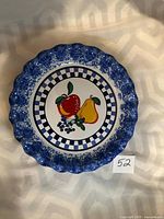 Top view of decorative pie plate showing scalloped blue rim, blue and white checkered border, and central fruit design of apple, pear, and blueberries.