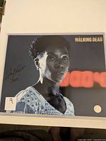 Color 8x10 photo of Jeryl Prescott signed in the left area with 'Walking Dead' logo in upper right corner. The photo is protected in a clear hard cover sleeve.
