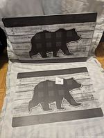 Two placemats laid flat showing rustic wood background with dark plaid bear silhouette design.