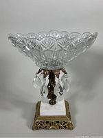 Side angled view of clear scalloped pressed crystal bowl with faceted drops hanging from brass mount, supported on white marble square base with ornate brass bottom