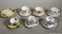 Full shot of 5 assorted bone china teacups, saucers, and dessert plates with various floral patterns and colors, arranged on flat surface.
