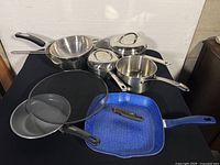 Overview of the cookware set showing various stainless steel pots and pans with and without lids, metal sieve, and blue enamel cast iron griddle pan.