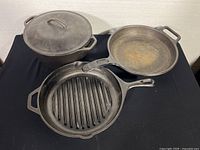 Three Lodge cast iron cookware pieces arranged together on black cloth: large covered Dutch oven, grill skillet, and frying pan showing wear and rust spots.