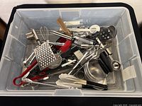 Large bin filled with assorted kitchen utensils, showing spatulas, graters, whisks, ladles, tongs, and more metal and plastic tools.
