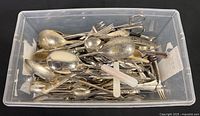 Top-down view of a plastic bin filled with assorted vintage metal spoons, forks, and other kitchen utensils