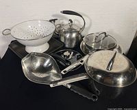 Photo showing vintage stainless steel whistling stovetop tea kettle, white enamel colander, stainless steel oval colander with black handle, square cast aluminum grill pan, and stainless steel pots with lids.