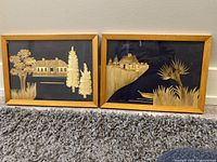 Two straw art pieces side by side on carpet floor showing rural house and nature scenes.
