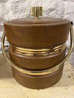 Overall front view of the ice bucket on a stone surface showing the brown faux leather exterior, brass band, curved brass handle, and brass knob on lid.