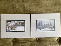 Two framed art prints by Loren Chabot with white mats, showing urban winter scenes side by side on a brown surface.