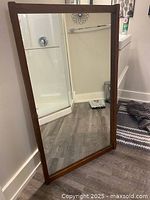 Front view of the large rectangular mirror leaning against a wall, showing the teak wood frame and reflective glass reflecting a shower area and other bathroom items.