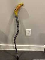 Full view of the painted folk art cane standing against a gray wall, showing its curved handle and decorated shaft.