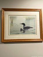 View of framed painting of a loon and chick floating on water with reeds and lily pads, signed and numbered.
