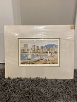 Full view of the artwork and mat board in plastic protective sleeve showing city skyline, beach, and sailboats on the water.