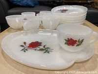 Top view showing seven white milk glass coffee cups and seven snack trays laid out on a table. Each cup and tray features red rose decals in excellent condition with no visible damage.