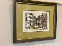 Framed watercolour painting of an urban street scene with buildings and a vehicle, showing artist's signature and details of frame and mat