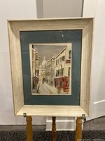 Framed vintage lithograph showing a narrow street with buildings and a person, mounted and matted under glass.
